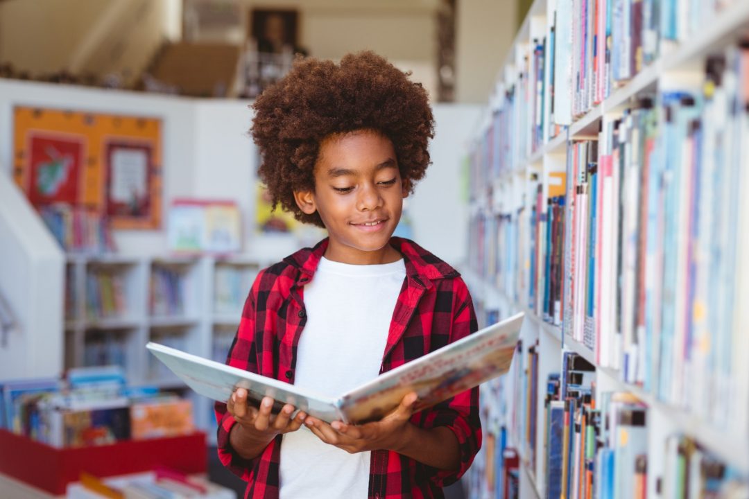smiling african american schoolboy reading book standing in school library