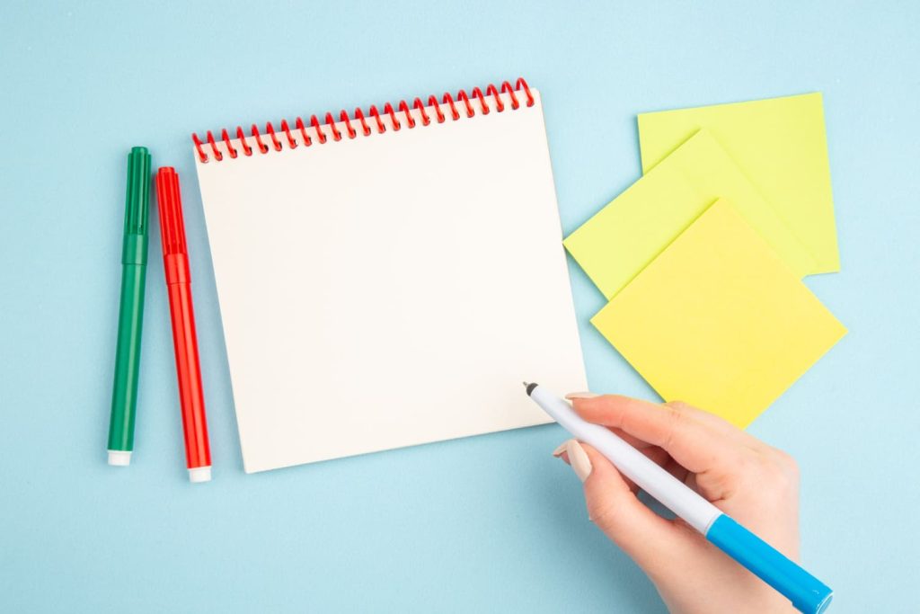 Top view of pen in hand with notepad and yellow sticky papers.